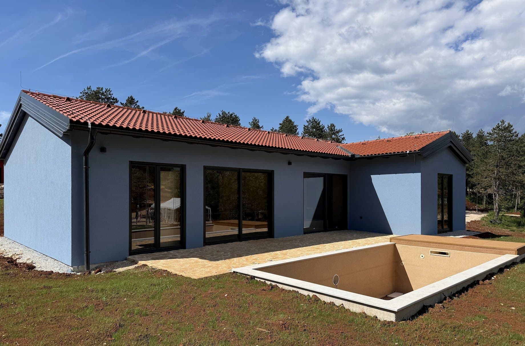 A completed single-storey home with a stone terrace and a new pool, pines behind it